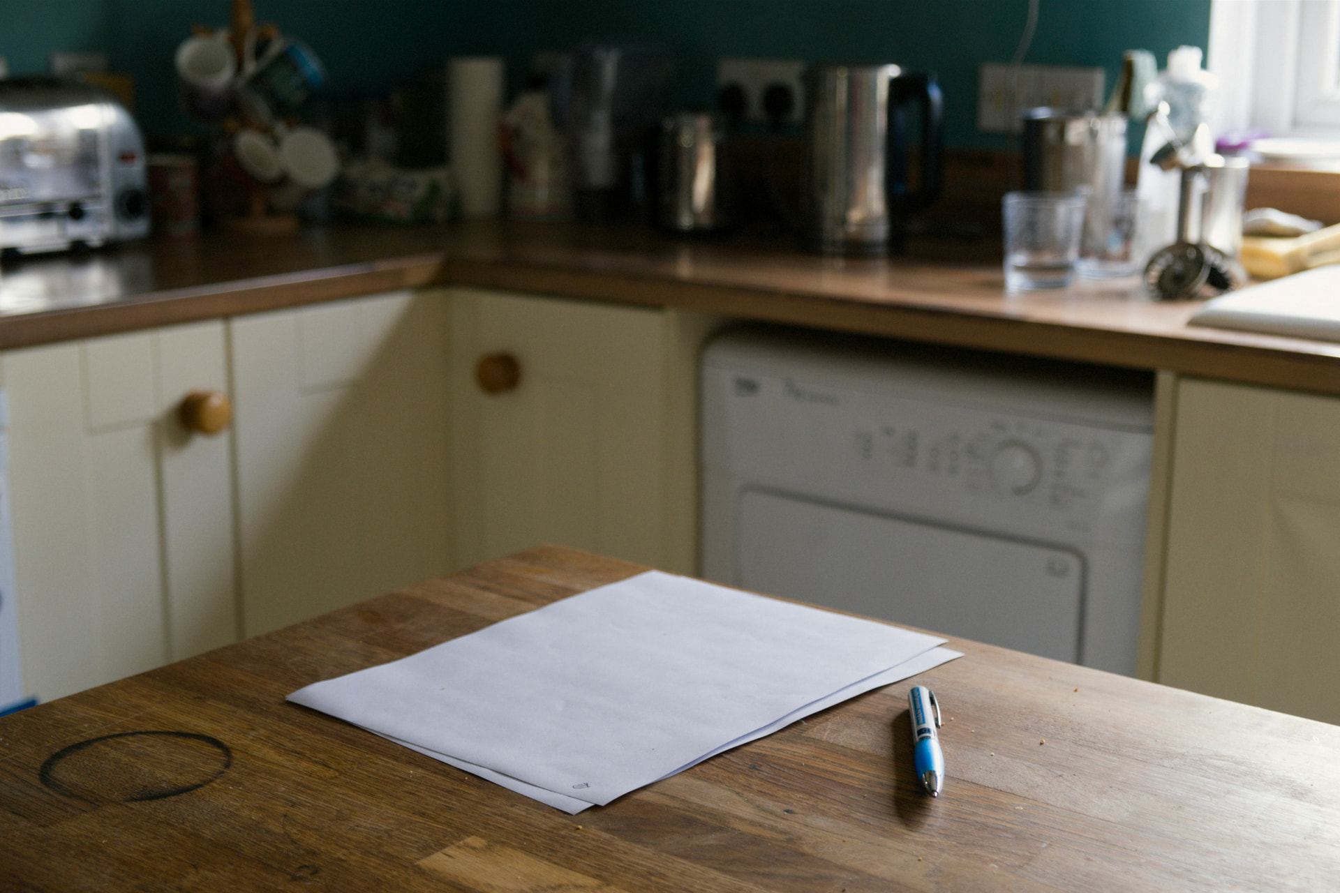 A blank sheet of white paper and a blue ballpoint pen resting on a wooden kitchen table, with softly blurred kitchen counters and cabinets in the background