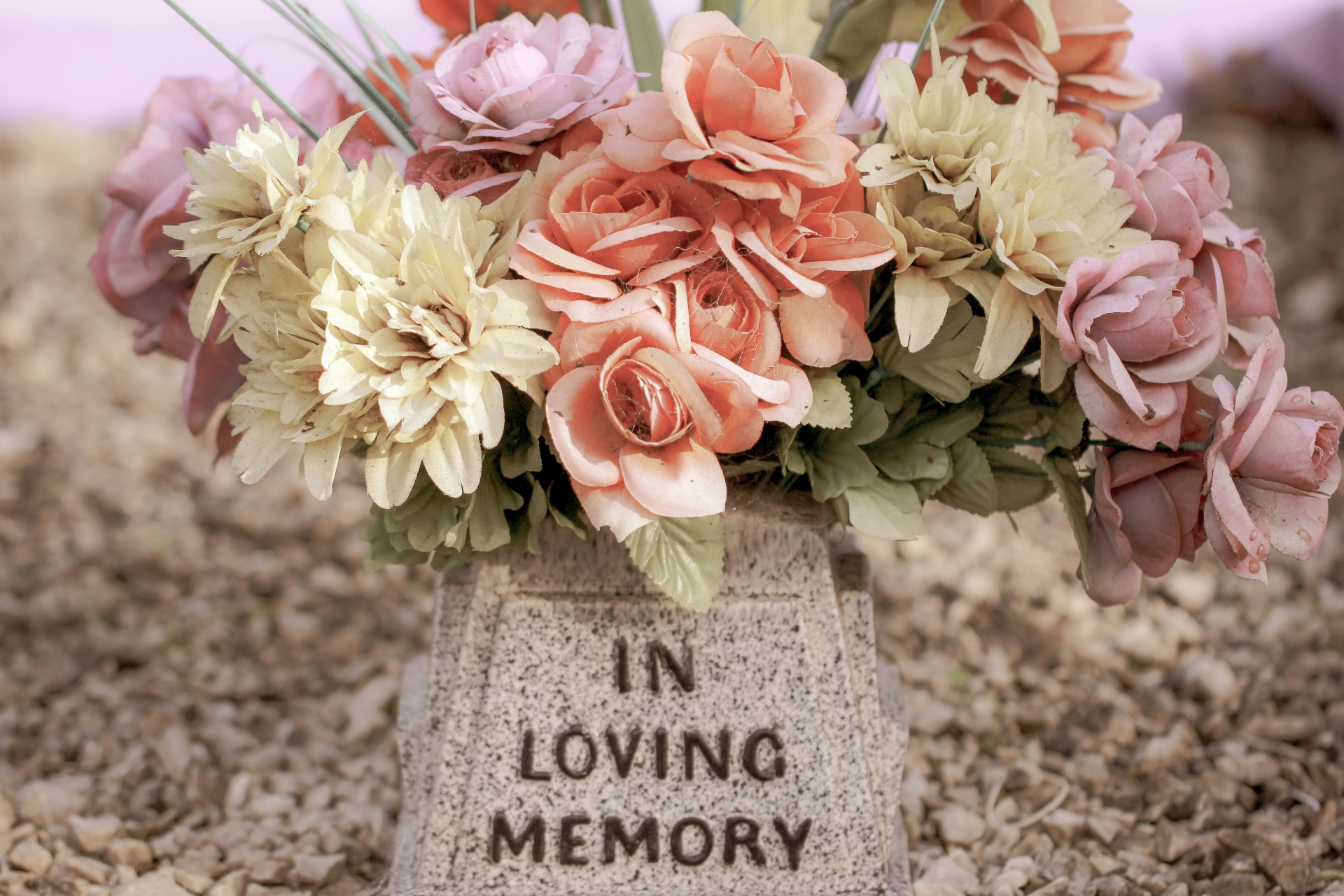 Pink and peach flowers arranged in an 'In Loving Memory' memorial vase on a bed of gravel
