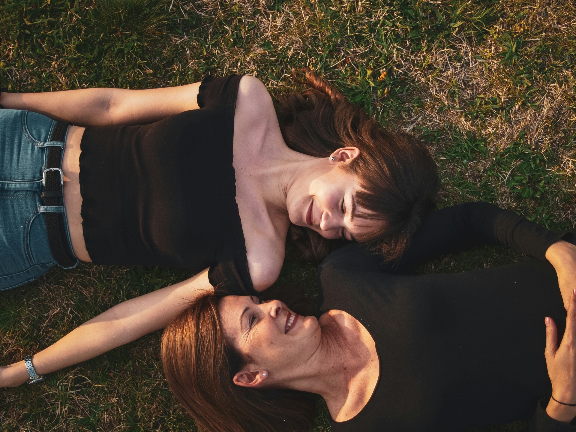 A mother and her adult daughter lying head to head on green grass, smiling in golden sunlight