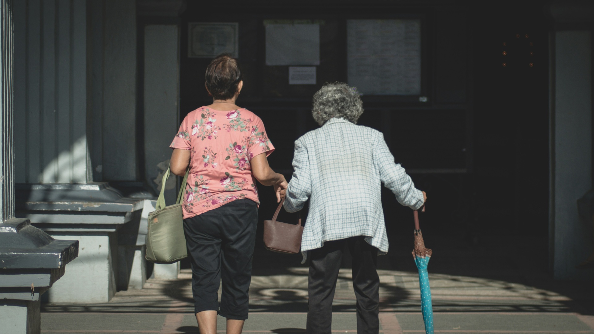A caregiver walking arm-in-arm with an elderly woman holding a cane outside a building